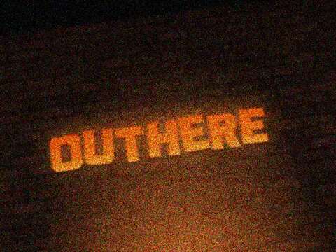 The orange cut-out OUTHERE sign on the brick, lit from below, photographed at an angle at night.