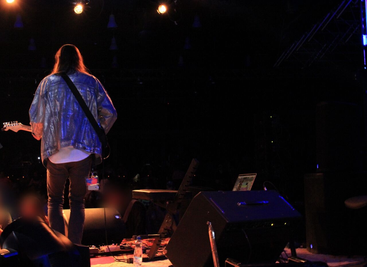 From the side of the stage: a guitarist in a denim jacket, back to the camera, monitors and a pedalboard at their feet, the room dark beyond.