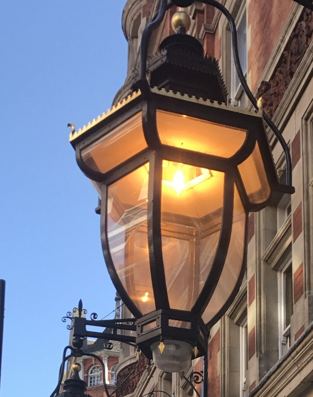 A big lit lantern on an iron bracket against a blue sky, red brick behind.